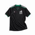 Maglia third Messico Coppa del Mondo 2026 — vista frontale su superficie in legno, colore nero con pattern geometrico azteco tono su tono, colletto rotondo con bordino tricolore verde-bianco-rosso e stemma nazionale sul petto sinistro