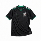 Maglia third Messico Coppa del Mondo 2026 — vista frontale su superficie in legno, colore nero con pattern geometrico azteco tono su tono, colletto rotondo con bordino tricolore verde-bianco-rosso e stemma nazionale sul petto sinistro